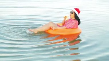 Bir yüzme daire ve Noel Baba şapka bir esmer kadın denizde Noel ve Yeni Yıl tutar bir muz yiyor