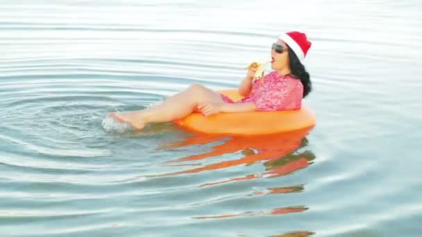 Uma mulher morena em um círculo de natação e um chapéu de Papai Noel come uma banana mantém o Natal e o Ano Novo no mar — Vídeo de Stock