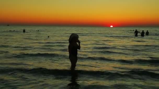 Žena v klobouku při západu slunce kráčí po pobřeží. Silueta — Stock video
