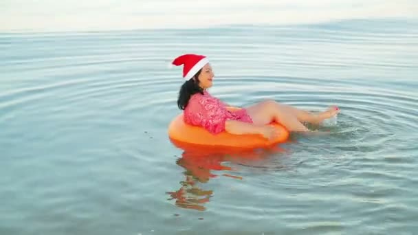 Egy barna nő egy úszó körben és egy Mikulás kalap tölti a karácsonyt és az újévet a tengeren — Stock videók