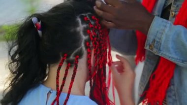 Koyu tenli bir kız, kızıl saçlı esmer bir kadına Afrika örgüsü örer.
