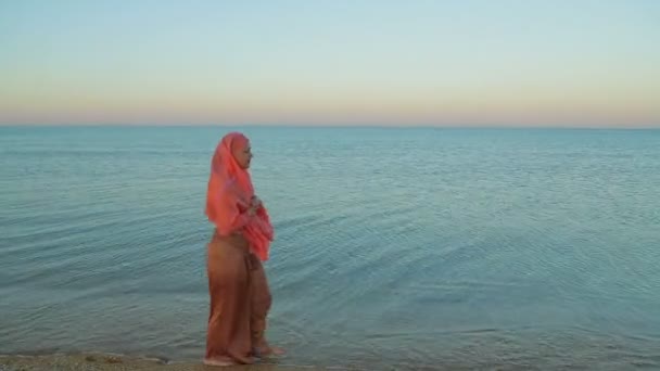 A young Muslim woman in an orange shawl walks along the seashore — Stock Video