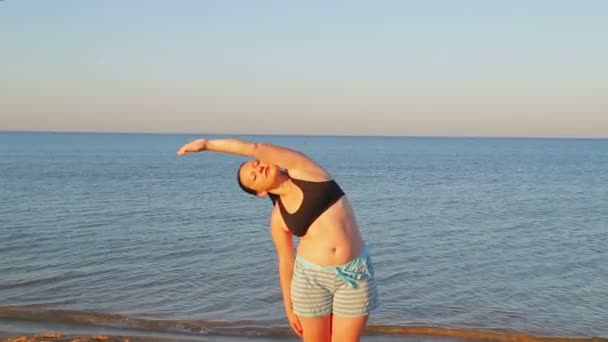 En brunett kvinna i shorts gör stretchingövningar på stranden — Stockvideo