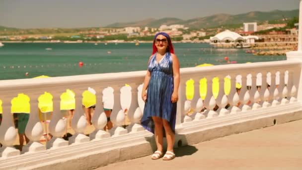 Eine Frau im blauen Kleid steht auf der Promenade und zeigt auf das Meer. — Stockvideo