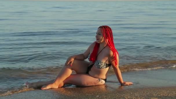 Женщина в купальнике сидит на берегу моря, по которому бегут волны — стоковое видео