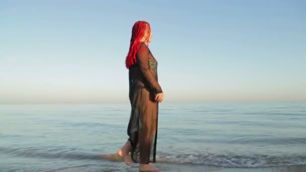 Een vrouw in een badpak en een zwart gewaad op de zee wandelingen langs de kust — Stockvideo