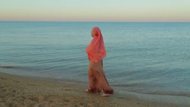 A young Muslim woman in an orange shawl walks barefoot along the seashore. — Stock Video