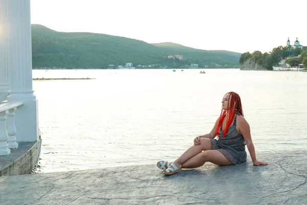 Hermosa mujer con el pelo rojo se sienta junto al lago en el fondo de las montañas al atardecer . — Foto de Stock