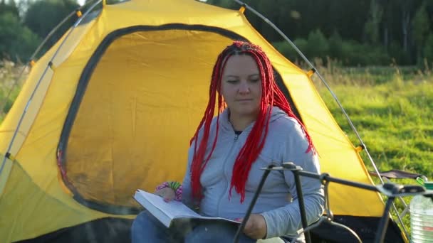 Una donna con le trecce africane vicino a una tenda da campeggio legge e guarda una pentola bollente — Video Stock
