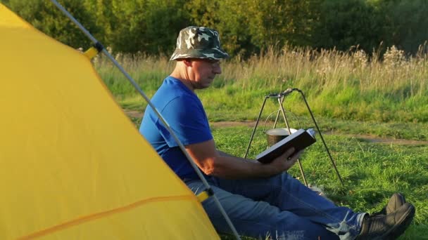 Een man in de buurt van een kampvuur tent leest en kijkt een kokend potje. — Stockvideo