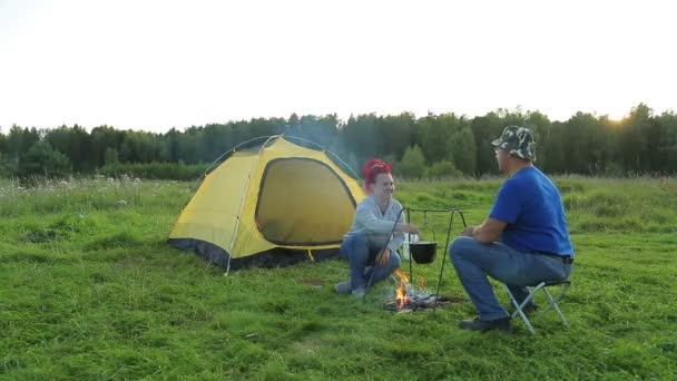 Een man en een vrouw in de avond in de buurt van een tent in de buurt van een vuur maken diner in een pot. — Stockvideo