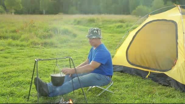 Een man in de buurt van een tent bij het vuur en horloges een kookpot. — Stockvideo