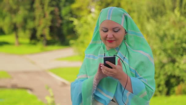 公園の軽いスカーフを持つ若いイスラム教徒の女性は、コーヒーを飲んで、ヘッドフォンでメッセンジャーで話しています — ストック動画