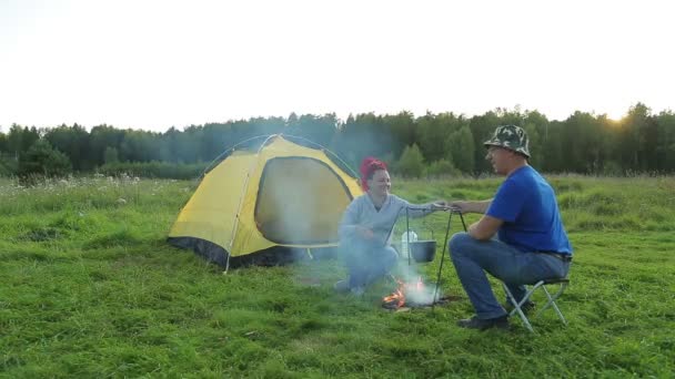 En man och en kvinna på kvällen nära ett tält nära en brand gör middag i en kruka. — Stockvideo