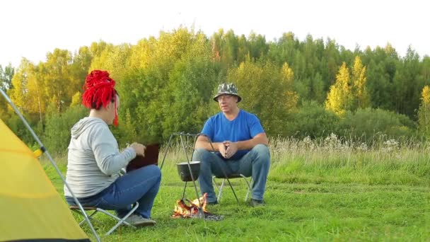 Een man en een vrouw in een clearing in de buurt van een tent in de buurt van een vuur maken diner in een pot — Stockvideo