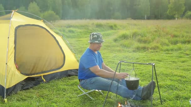 Um homem perto de uma tenda perto do fogo e observa uma panela fervente . — Vídeo de Stock