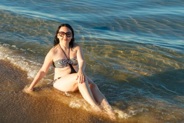 Deniz kıyısında mayo ve gözlüklü esmer bir kadın oturuyor..