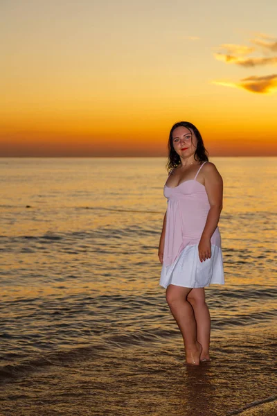 Молодая женщина в белом платье стоит на берегу моря на закате. — стоковое фото