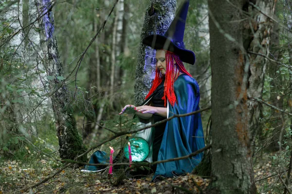 La hechicera con un sombrero y una capa negra en el bosque encantos sobre una bola de cristal luminoso. Fotografía horizontal — Foto de Stock