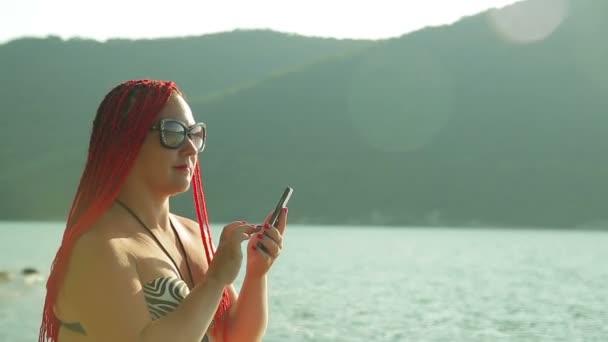Женщина с рыжими волосами ведет блог на берегу на фоне гор — стоковое видео