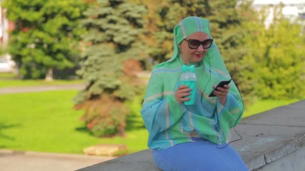 A young cheerful Muslim woman in a light scarf and sunglasses in the park drinks coffee and talks in the messenger with headphones. — Stock Video