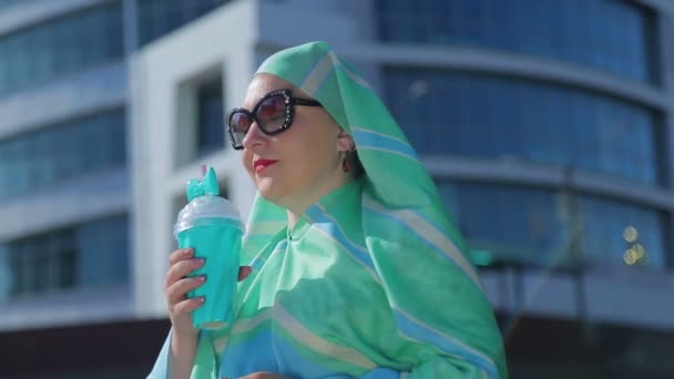 A young Muslim woman in a light scarf and sunglasses against the background of a modern building. — Stock Video