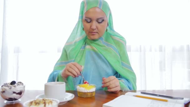 Een jonge moslim vrouw in een licht groene sjaal in een café eet taarten en drankjes thee. — Stockvideo