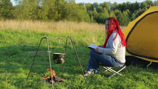 Žena poblíž šátek u ohně čte knihu a pozoruje vroucí hrnec. — Stock video