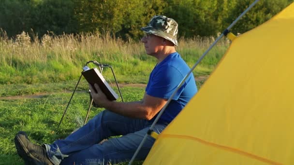 Um homem perto de uma tenda de fogueira lê e observa uma panela fervente . — Vídeo de Stock