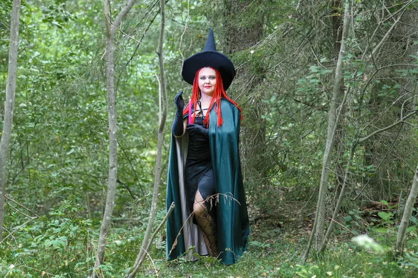 Una hechicera con el pelo rojo en un sombrero puntiagudo y una capa negra en el bosque se dedica a los encantos con una vela . — Foto de Stock