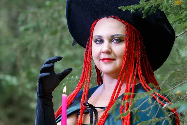 Close-up Sorceress with red hair in a hat and a black cloak in the woods in the hand with a candle. — Stock Photo, Image