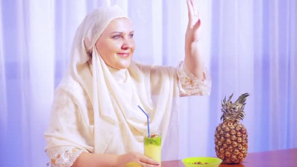 En ung muslimsk kvinna i en lätt halsduk dricker en fruktsmoothie och ler — Stockvideo