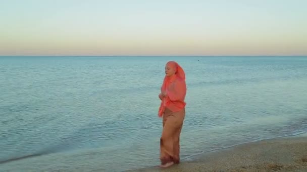 Молодая мусульманка в оранжевой шале ходит босиком по берегу моря — стоковое видео