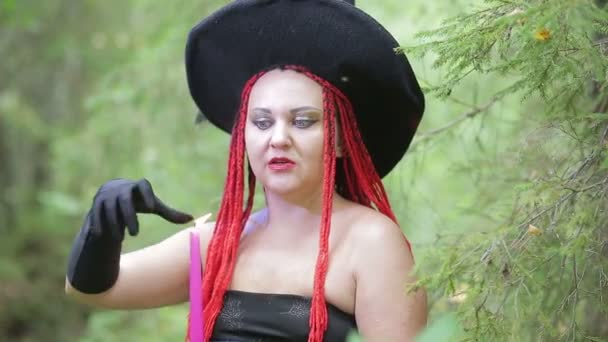 Bruja joven seductora con el pelo rojo y un sombrero puntiagudo evoca con velas en el bosque — Vídeo de stock