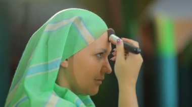 Ayna nın önünde müslüman bir kadın fırçayla bir fondötenle makyaj yapar.