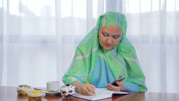 Een jonge moslim vrouw in een licht groene sjaal in een café werkt op afstand met een telefoon en een dagboek. — Stockvideo