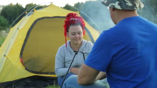 Een man en een vrouw in een clearing in de buurt van een tent in de buurt van een vuur maken diner in een pot. — Stockvideo