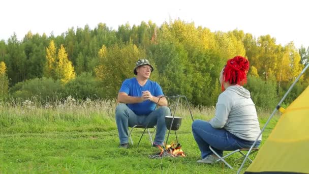 Een man en een vrouw in een clearing in de buurt van een tent in de buurt van een vuur maken diner in een pot — Stockvideo