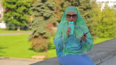 Parkta hafif bir eşarp ve güneş gözlüğü genç bir neşeli Müslüman kadın kahve içiyor ve kulaklık ile haberci konuşuyor.