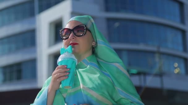Eine junge muslimische Frau mit hellem Schal und Sonnenbrille vor dem Hintergrund eines modernen Gebäudes — Stockvideo