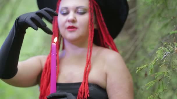 Una bruja joven con el pelo rojo y un sombrero negro hace magia en el bosque — Vídeo de stock