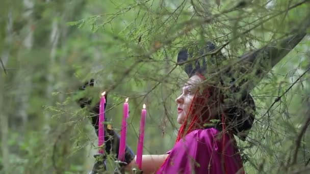 Eine junge Hexe mit roten Haaren und schwarzem Hut und Himbeermantel übt im Wald Magie. — Stockvideo