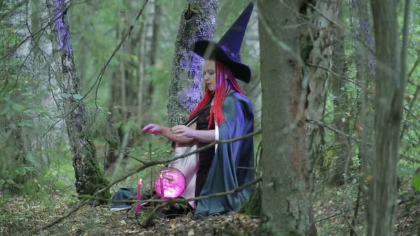 Uma jovem bruxa com cabelo vermelho e um chapéu preto e capa preta está envolvida em magia na floresta — Vídeo de Stock