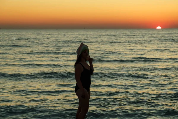 Una mujer con sombrero camina a lo largo de la orilla del mar al atardecer . — Foto de Stock