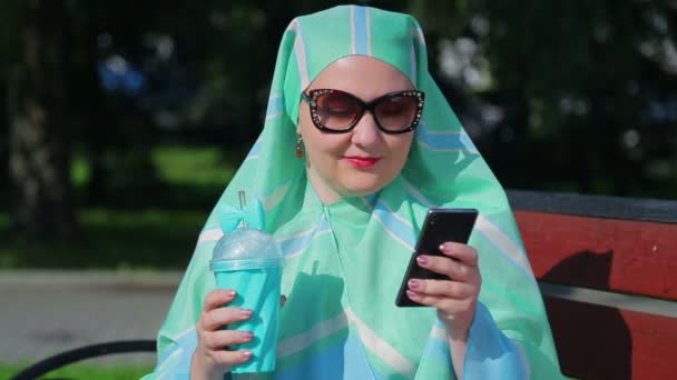 Una giovane donna musulmana in una sciarpa leggera e occhiali scuri siede su una panchina nel parco e beve caffè e parla nel messaggero. . — Video Stock
