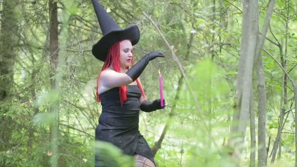 Bruja joven seductora con el pelo rojo y un sombrero puntiagudo evoca con velas en el bosque . — Vídeos de Stock
