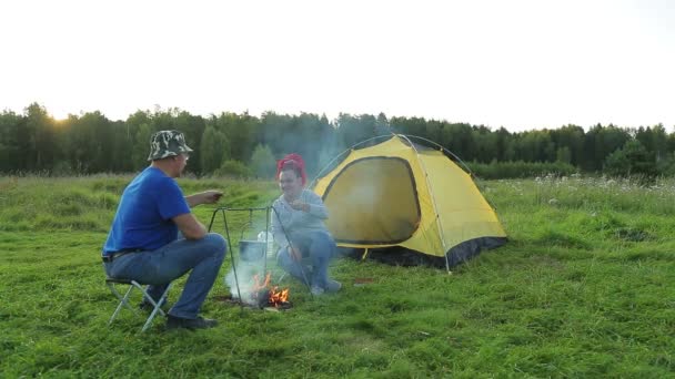 A man and a woman in the evening near a tent near a fire make dinner in a pot. — Stock Video