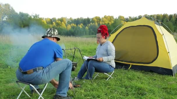 Muž a žena při západu slunce u stanu nedaleko ohně dělají večeři v hrnci. — Stock video