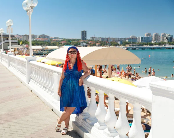 Una mujer con coletas africanas en un vestido azul se levanta en el paseo marítimo sobre el fondo del mar . — Foto de Stock