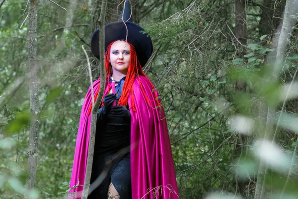 A sorceress with red hair in a hat and a purple cloak in the forest in her hand with a burning candle. — Stock Photo, Image